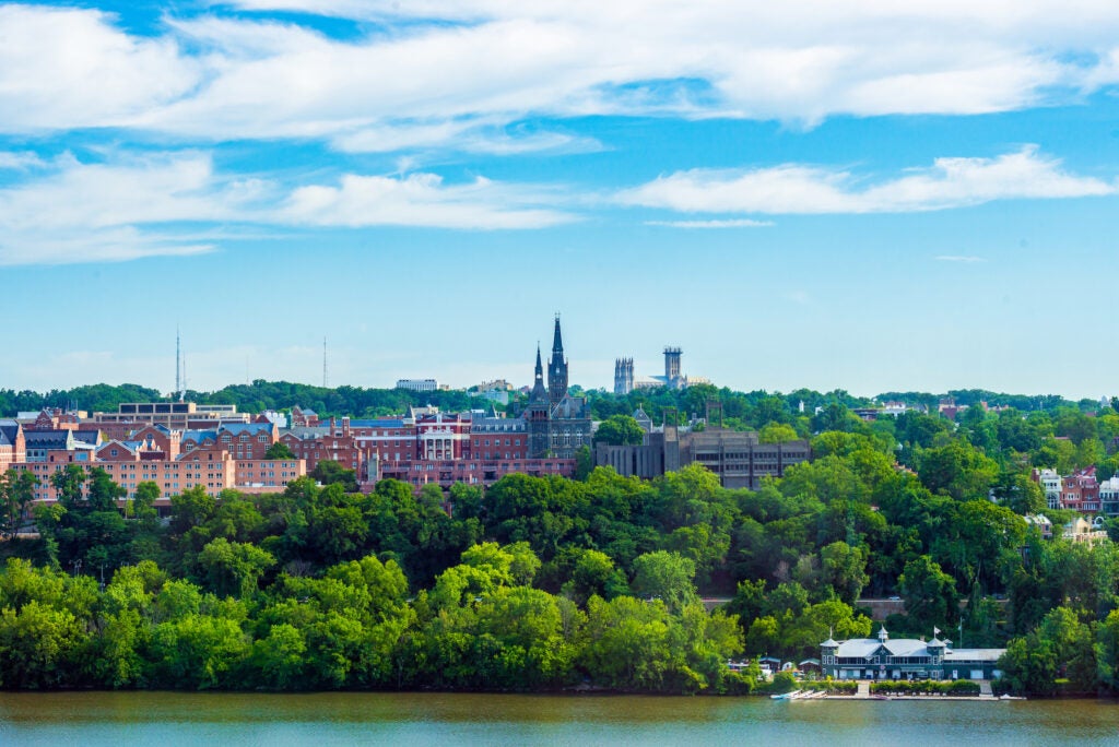 Image: A skyline view of the Georgetown University campus, slightly cloudy blue sky, and full green trees.